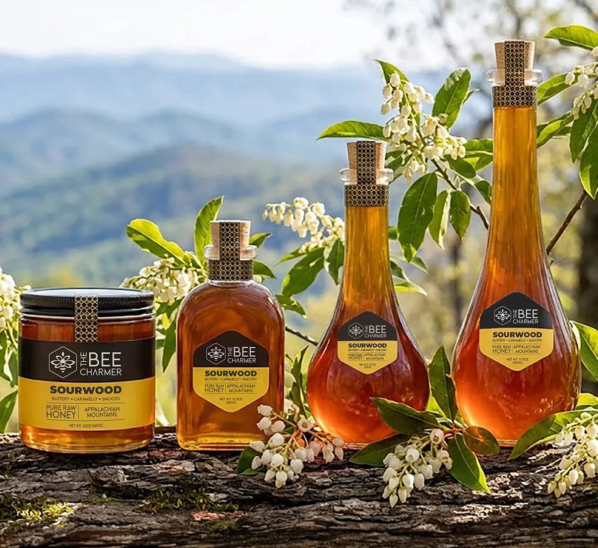 Four honey products from The Bee Charmer arranged on a log outdoors with green leaves and white blossoms against a blurred mountain landscape. The products include an assortment of different size jars and bottles of sourwood honey.