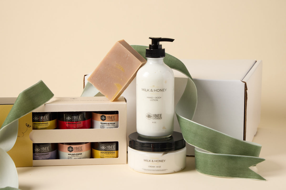 The Bee Charmer Collection Gift Set: six jars of raw honey in a gift box with Milk and Honey bar soap, lotion and cream, shown with a green ribbon beside a white shipping box